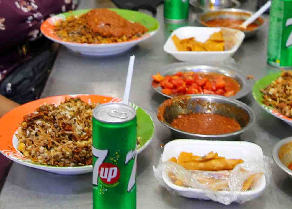 Close-up view of a meal featuring a can of 7up, plates of Koshari which is a dish with rice, pasta, tomatoes and chickpeas and various sauces and toppings on a table.