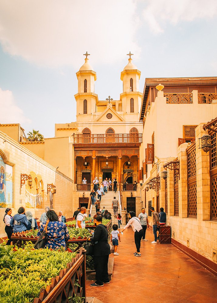 Courtyard of the Hanging Church in Cairo, Egypt, bustling with visitors and featuring twin bell towers in the background.