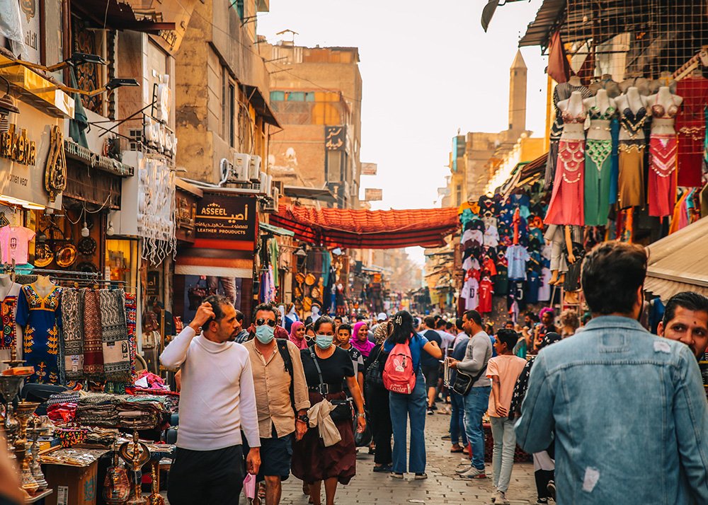 A bustling market street in the Khan el-Khalili bazaar filled with people shopping and various colorful textiles, clothing, and goods displayed in shops on either side.