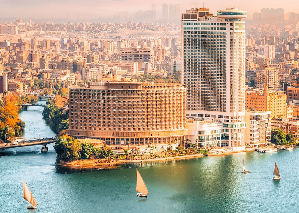 An aerial view of Cairo, showcasing the Nile River, tall buildings, and sailboats on the water.