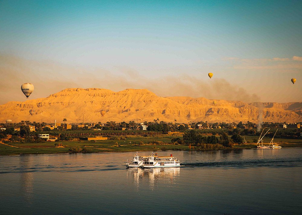 Hot air balloons floating over the Nile River with a cruise ship in the foreground and desert mountains in the background during sunrise.