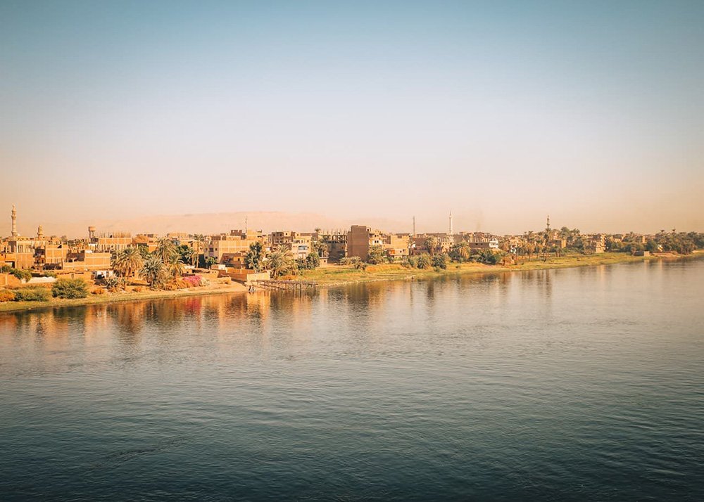 A view of Luxor's West Bank with buildings, palm trees, and greenery along the shore, under a clear blue sky