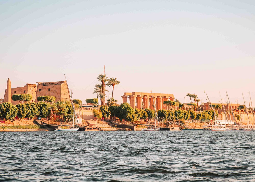 A view of Luxor Temple from across the river, showcasing ancient Egyptian architecture with palm trees and boats along the shoreline.