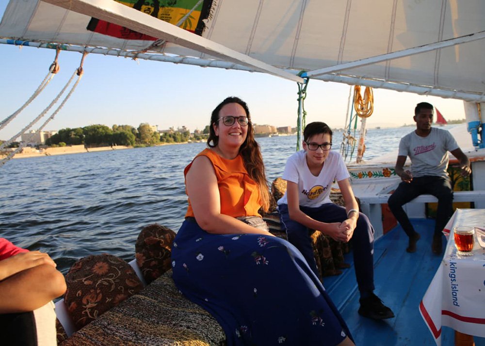 A woman and a boy sit in a felucca, an Egyptian sailing boat. In the background parts of the Luxor temples are visible.