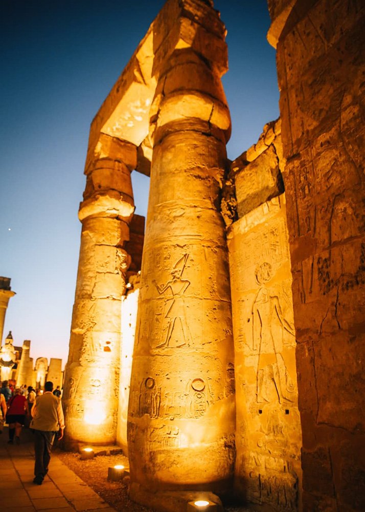 Ancient Egyptian pillars with hieroglyphs and carvings, lit warmly at night with people walking around.