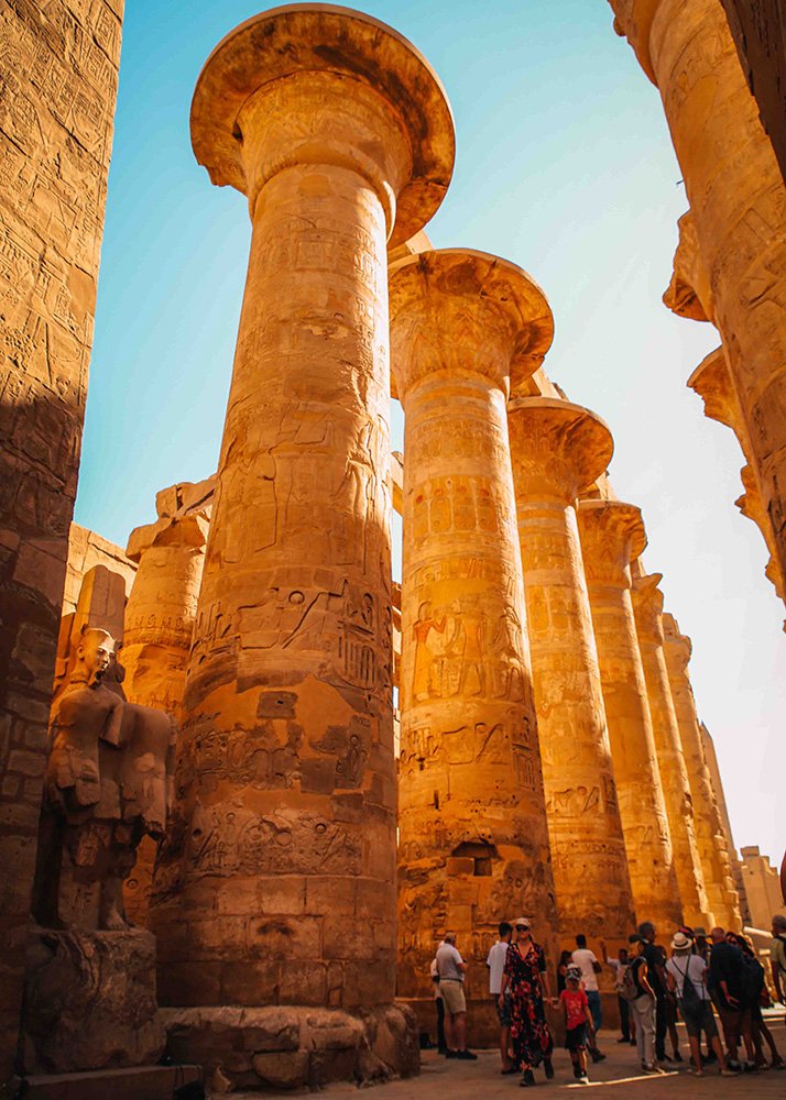 The towering columns of the Great Hypostyle Hall at Karnak Temple, covered in hieroglyphics and carvings, with visitors walking below.