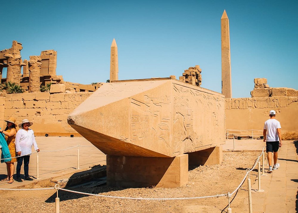 A large hieroglyphic block and several obelisks in an open-air historical site, Karnak Temple, with tourists observing the area