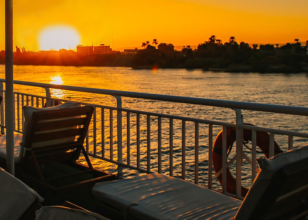 View from a Nile Cruise ship at sunset on the Nile River, with the sun setting behind the distant buildings and palm trees, casting a golden glow on the water.
