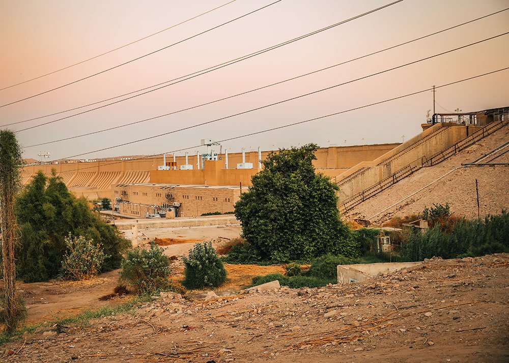 The Aswan High Dam at dusk with in front of it lush green trees and electric wiring cables can be seen hanging in the air.