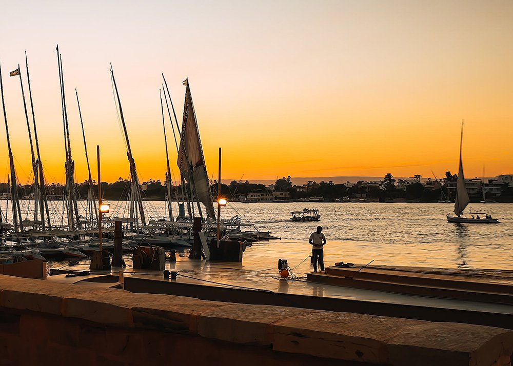 Feluccas docked along the Nile River at sunset, with their tall sails silhouetted against the glowing sky and a fisherman standing by the water.
