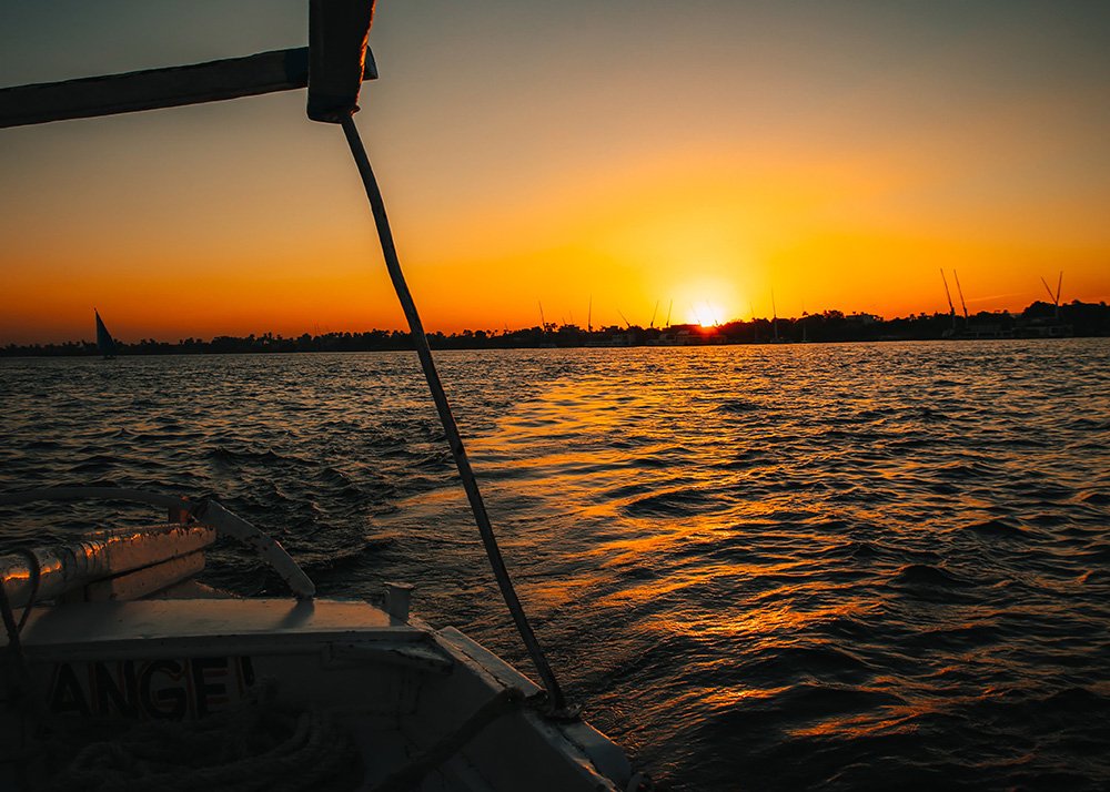 Sunset view from a felucca sailboat on the Nile, with the sky ablaze in orange hues and the silhouette of boats and palm trees on the horizon.