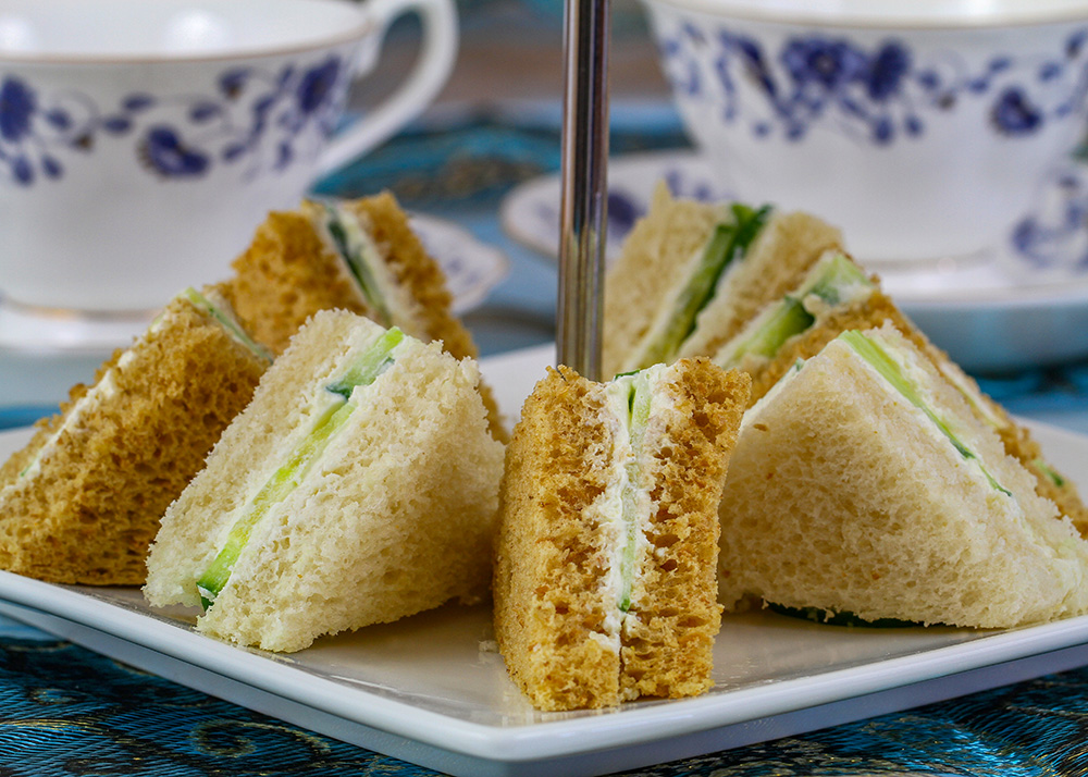 Finger sandwiches on a square white plate with 2 white and blue teacups behind it