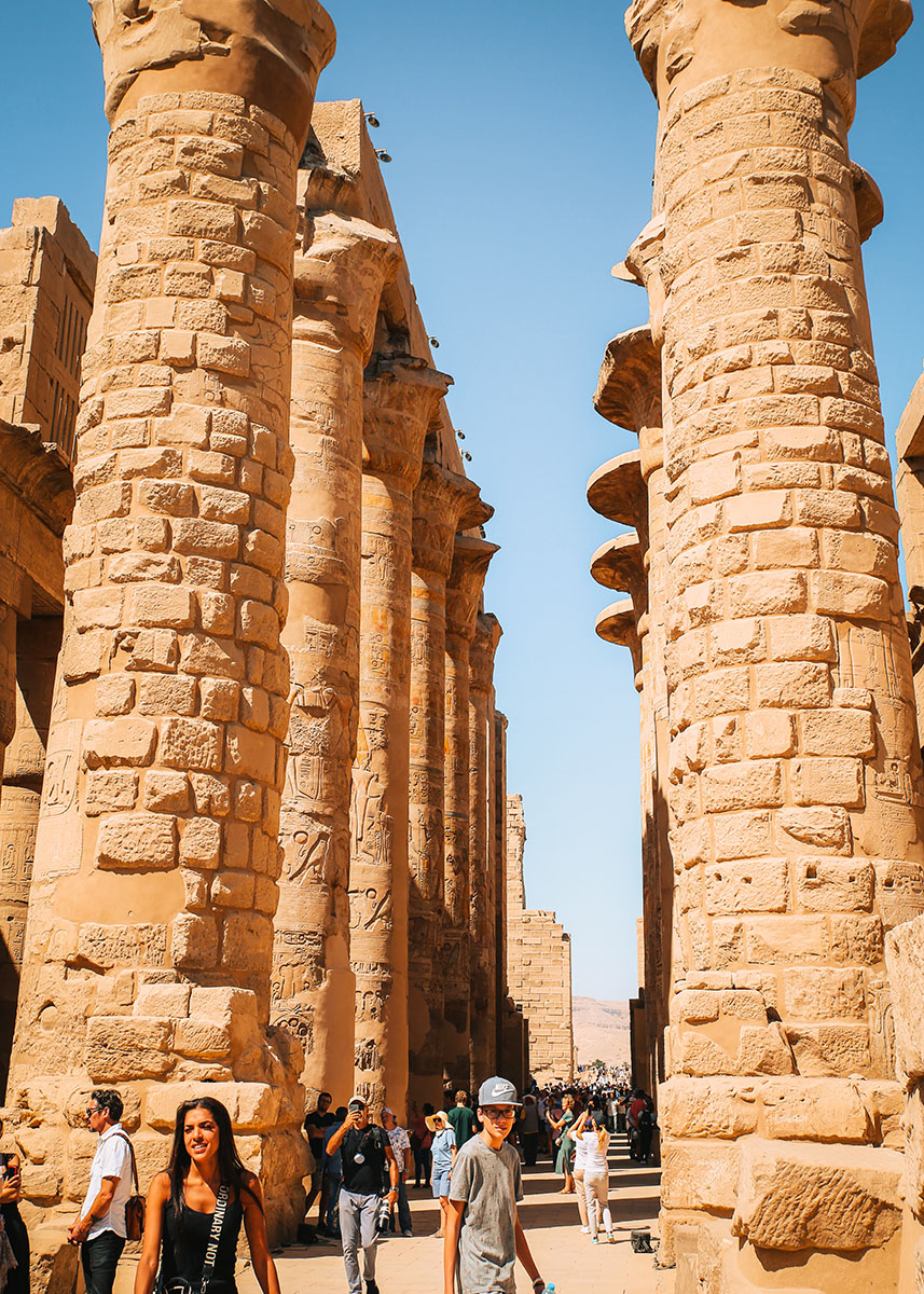 Visitors walk through towering stone columns covered in hieroglyphs at Karnak Temple in Luxor, Egypt, under a bright blue sky. A teenage boy in a cap and tourists with cameras explore the open-air ruins. The midday sun highlights the detailed carvings, offering a fun way to introduce kids to ancient history.