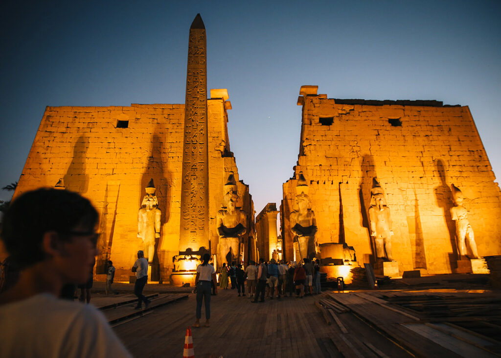 Crowds explore the grand entrance of Luxor Temple at dusk, with towering statues and an obelisk bathed in golden light. The ancient stone walls glow under the night sky as visitors move through the historic site. Ideal for kids to witness Egyptian grandeur lit up in dramatic contrast to the twilight.