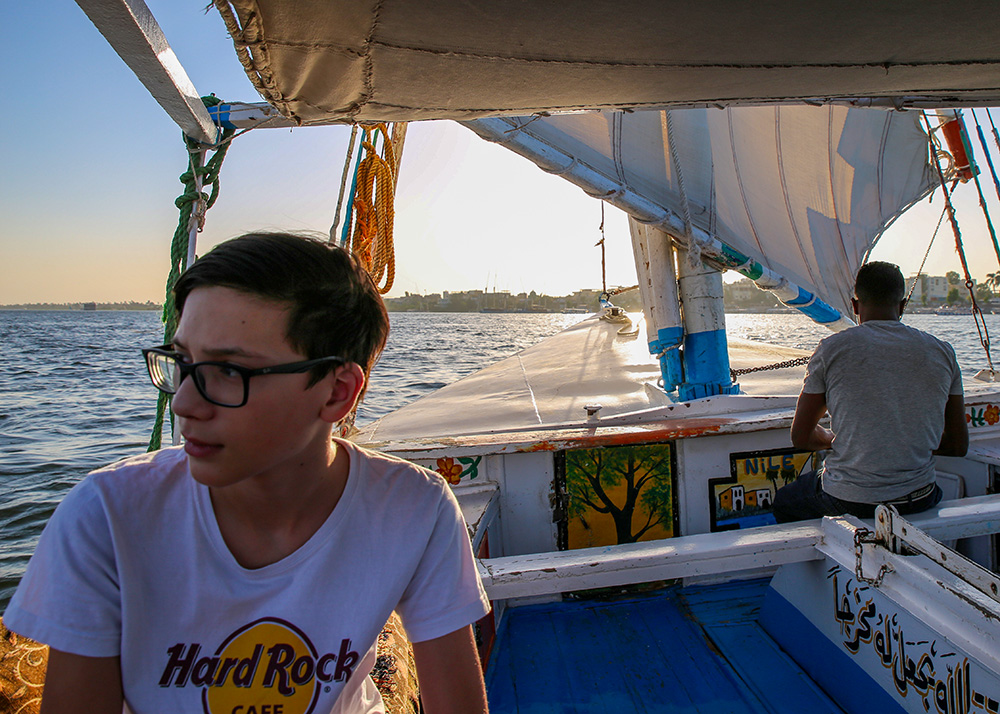A teenage boy is on a felucca on the Nile river wearing a white t-shirt