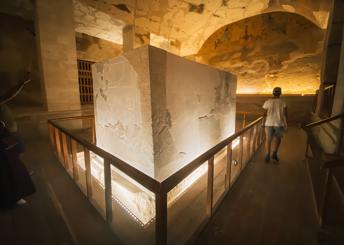A well-lit stone sarcophagus with faded hieroglyphics stands in the Tomb of Seti I, inside Luxor’s Valley of the Kings. The chamber has soft golden lighting and detailed wall paintings, while a casually dressed boy walks by. The atmosphere is quiet and reverent, perfect for educational exploration with kids.