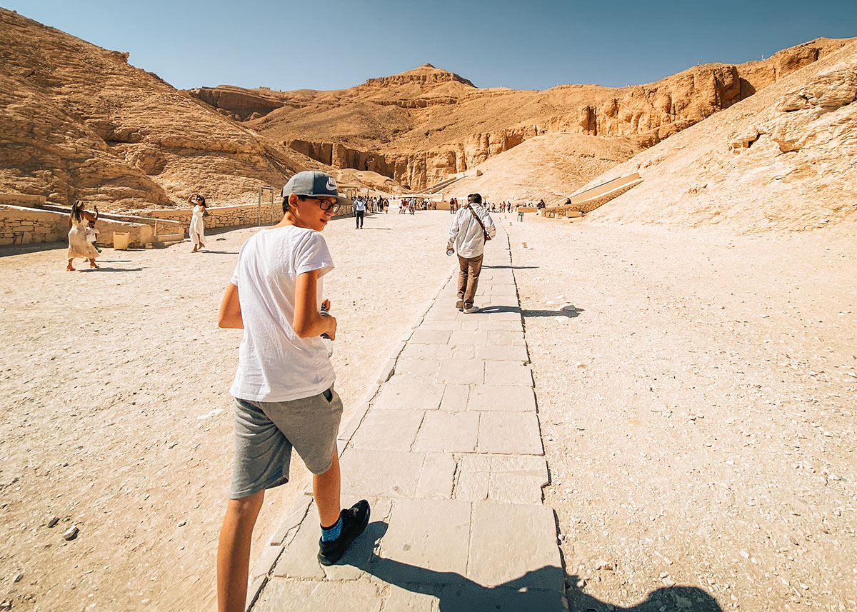 A teen wearing casual clothing and a cap walks on a walkway towards the tombs in the Valley of the Kings in Egypt