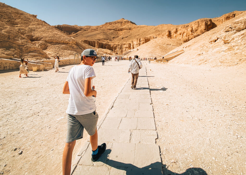 A teen wearing casual clothing and a cap walks on a walkway towards the tombs in the Valley of the Kings in Egypt