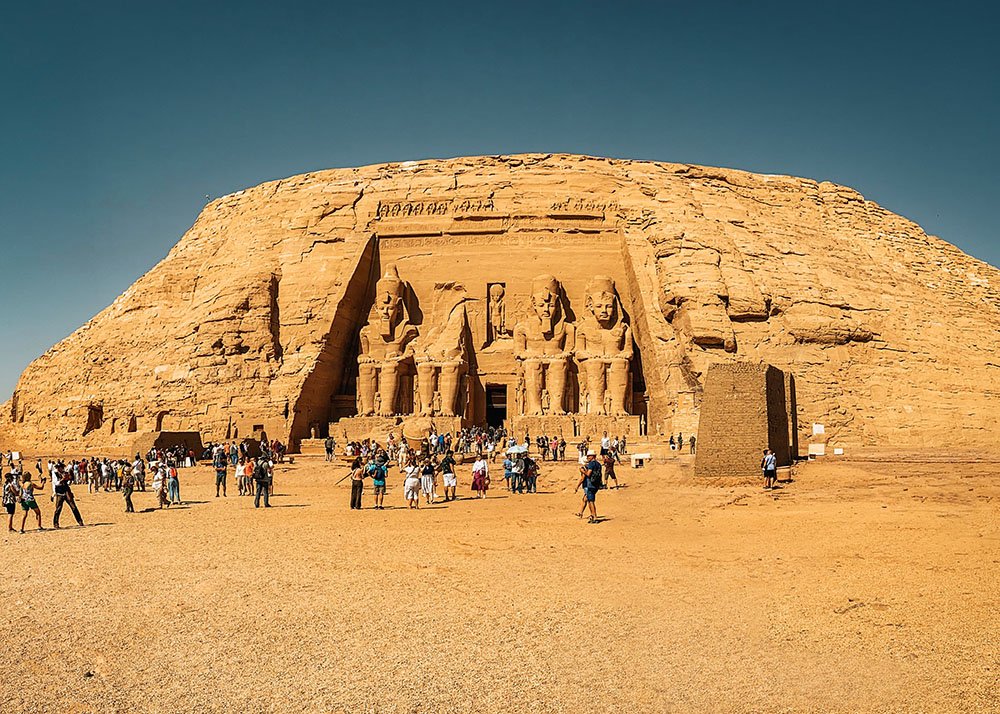 The grand facade of Abu Simbel with four colossal statues of Ramses II, surrounded by tourists, under a clear blue sky.