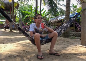 A boy sitting in a hammock, smiling to the camera and drinking a coke