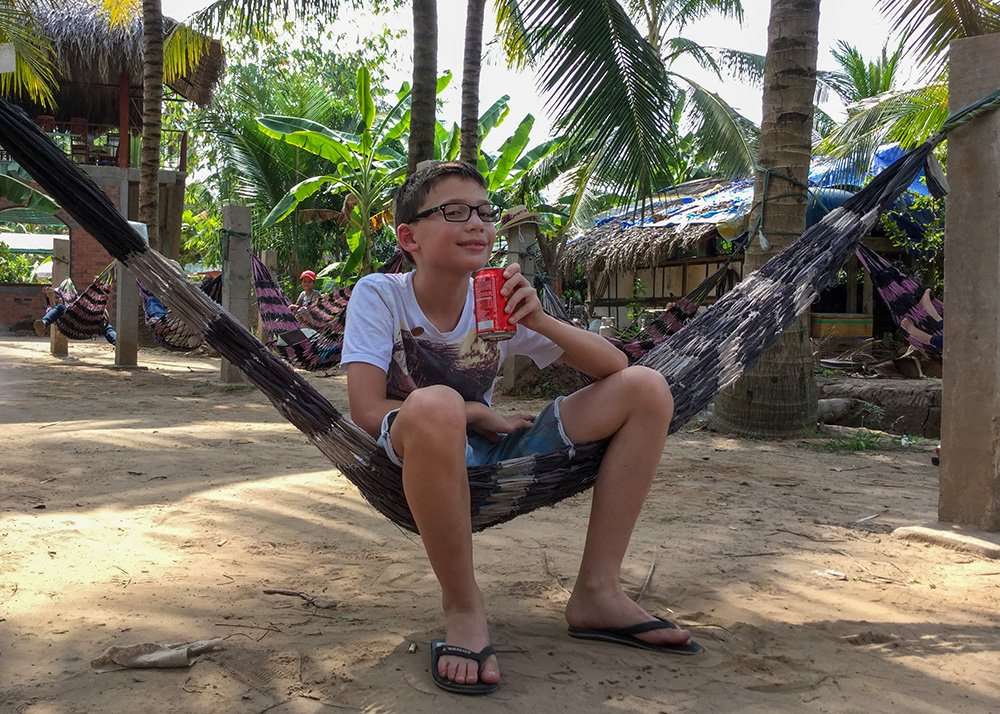 A boy sitting in a hammock, smiling to the camera and drinking a coke