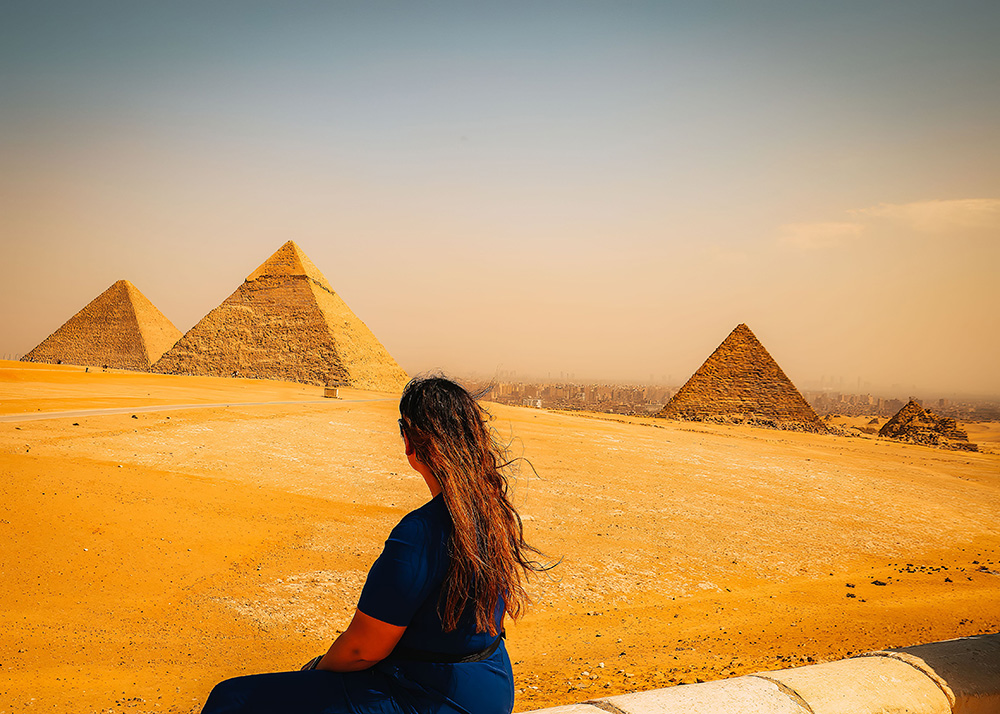 A woman with long brown hair and a blue jumpsuit sits on a ledge gazing at the pyramids of Giza in the distance, with the cityscape of Cairo faintly visible.