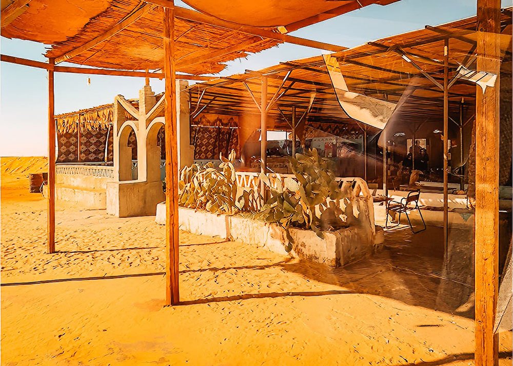 A rustic rest stop in the desert featuring shaded seating areas with traditional decor, surrounded by sand.