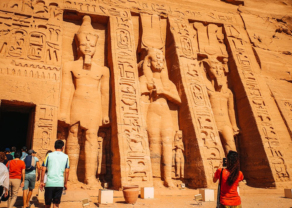 Tourists observing the right side of Ramses II's temple facade, which features colossal statues and intricate carvings.