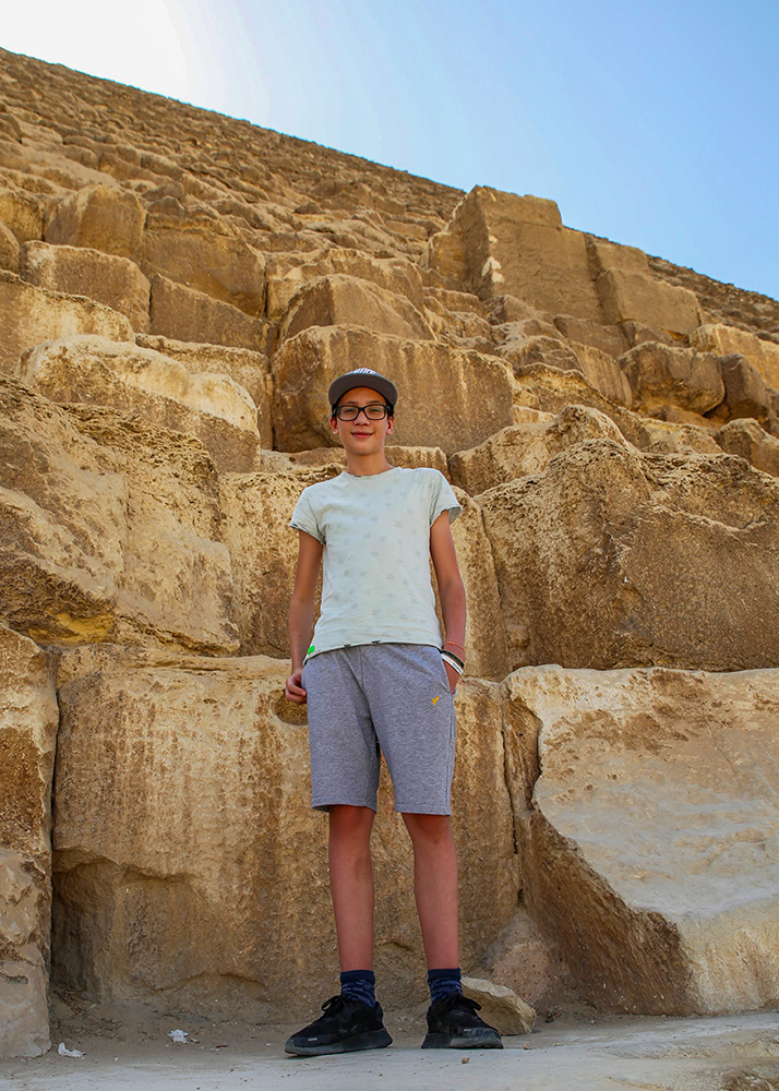 A boy with a cap, white shirt and short grey shirts is posing on the steps of one of the pyramids of Gizeh, looking down at the photographer with a proud look on his face.