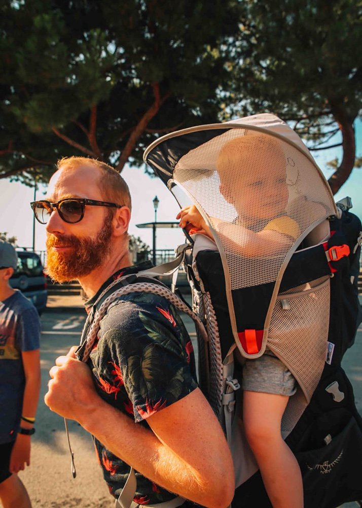 A bearded man with sunglasses carries a toddler in a backpack carrier, both looking content, with trees and a sunny sky in the background. It's a sunny day so the toddler is comfortable seated with a sunscreen on top of the back carrier