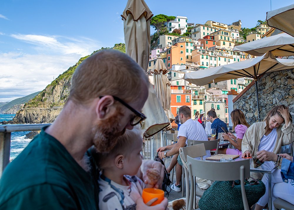 People dining at an outdoor café with the colorful houses and cliffs that Riomaggiore is known for in the background, exemplifying the charming and vibrant atmosphere of Cinque Terre.