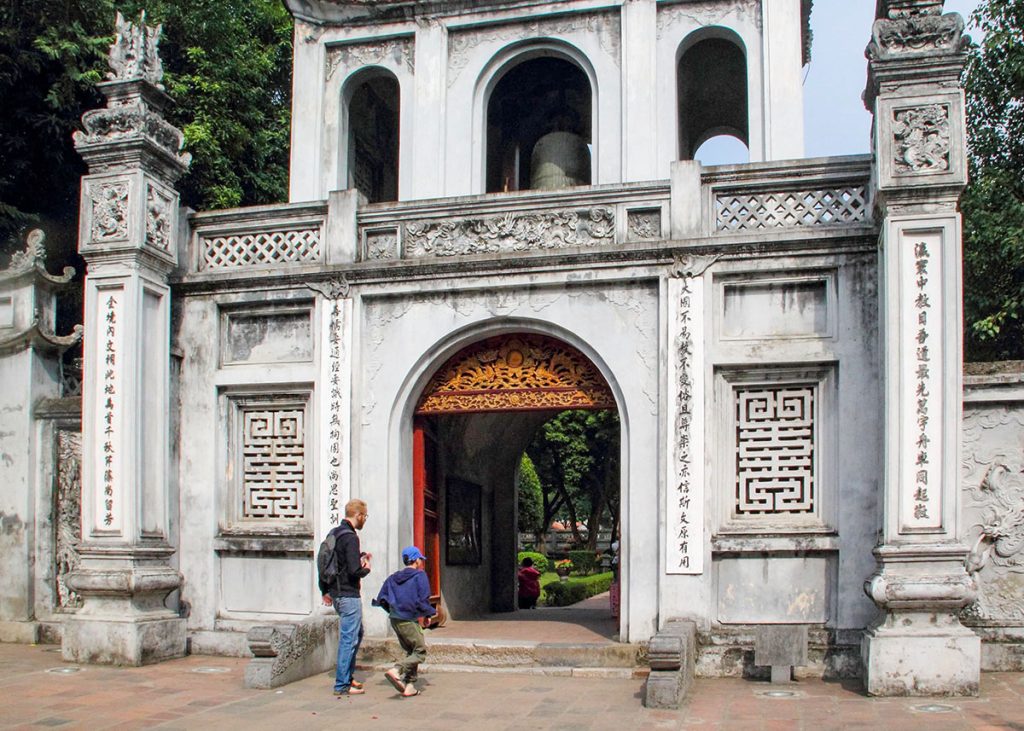 An adult and child walk toward the ornate entrance of the Temple of Literature in Hanoi, Vietnam, a historic Confucian temple and former university. The stone gate is decorated with traditional carvings and Vietnamese script, with greenery surrounding the site. The boy in a blue cap and hoodie appears curious as they approach, highlighting the temple’s appeal to both children and adults during a pleasant day out.