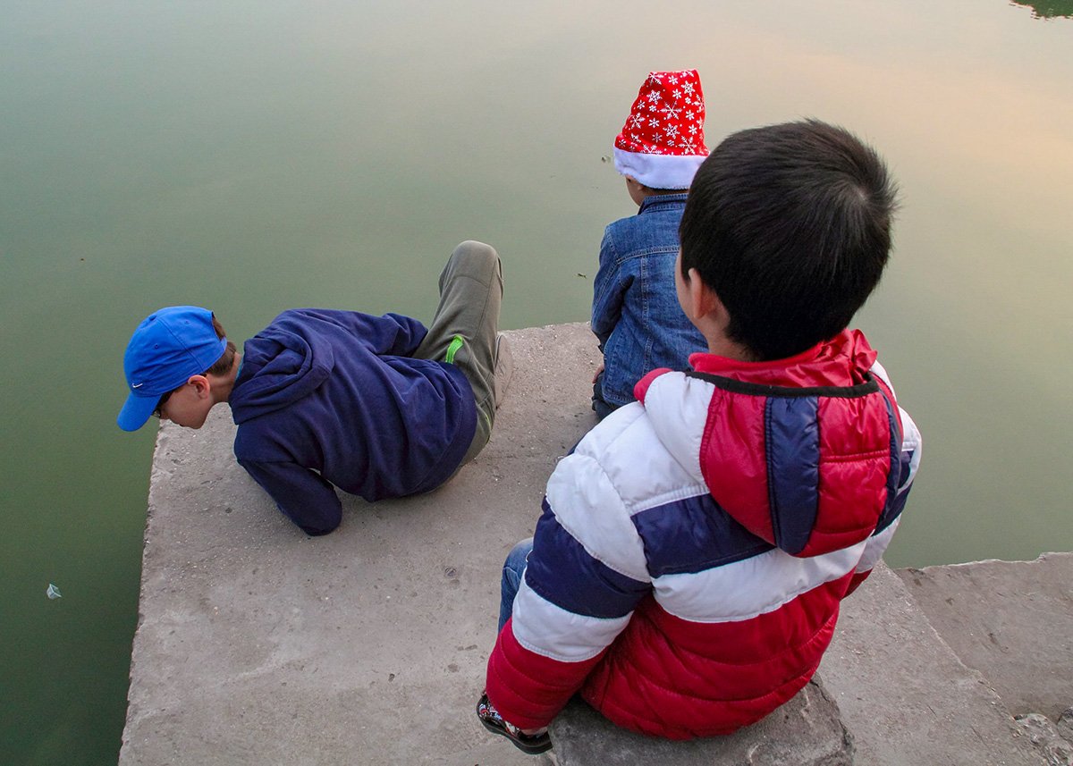 Three children sit on the edge of a concrete platform by Hoan Kiem Lake in Hanoi, Vietnam, with calm, greenish water reflecting the soft sunset light. One child leans over the edge curiously, possibly searching for the legendary Hoan Kiem turtle, while another wears a Santa hat, adding a playful holiday touch. The peaceful scene captures a quiet moment of childhood exploration in mild, clear weather.