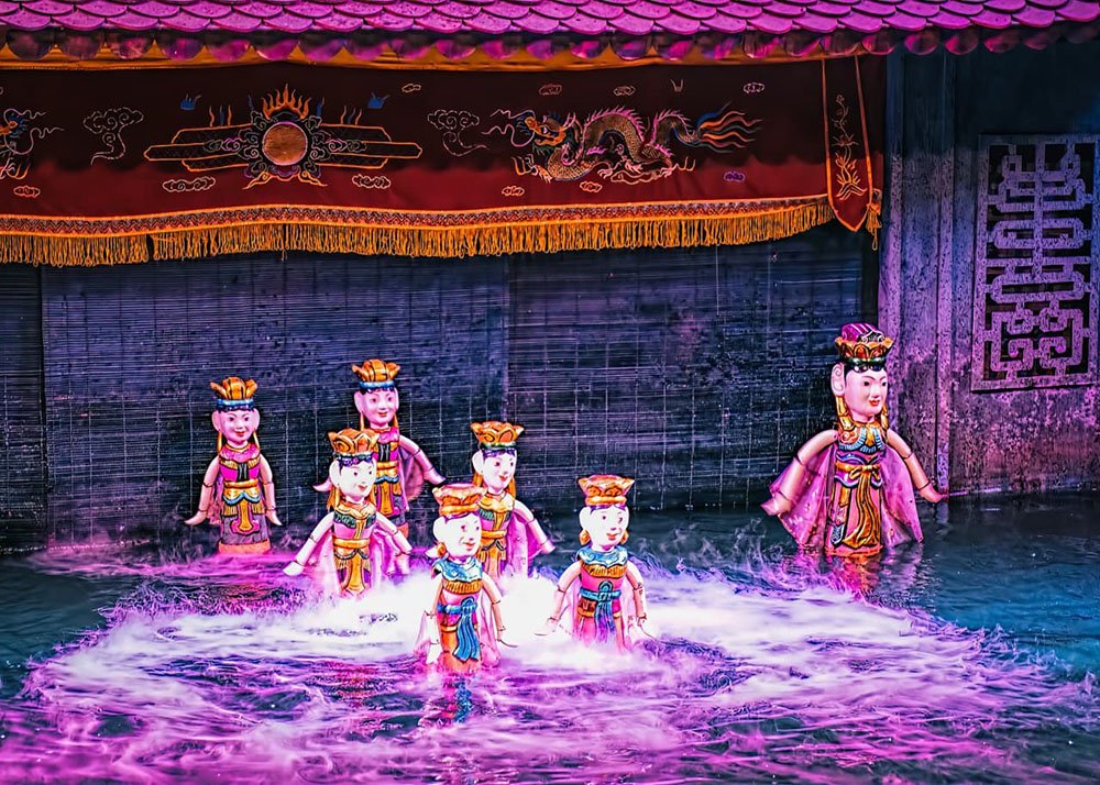 Wooden puppets wear handmade clothing and are dancing in the water puppet theatre. The lighting is purple which gives the scene a magical feel.