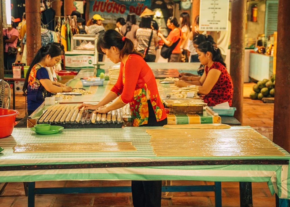Two woman working in a coconut candy factory. They're slicing big parts of the coconut candy into small pieces for consumption, while tourists look ong