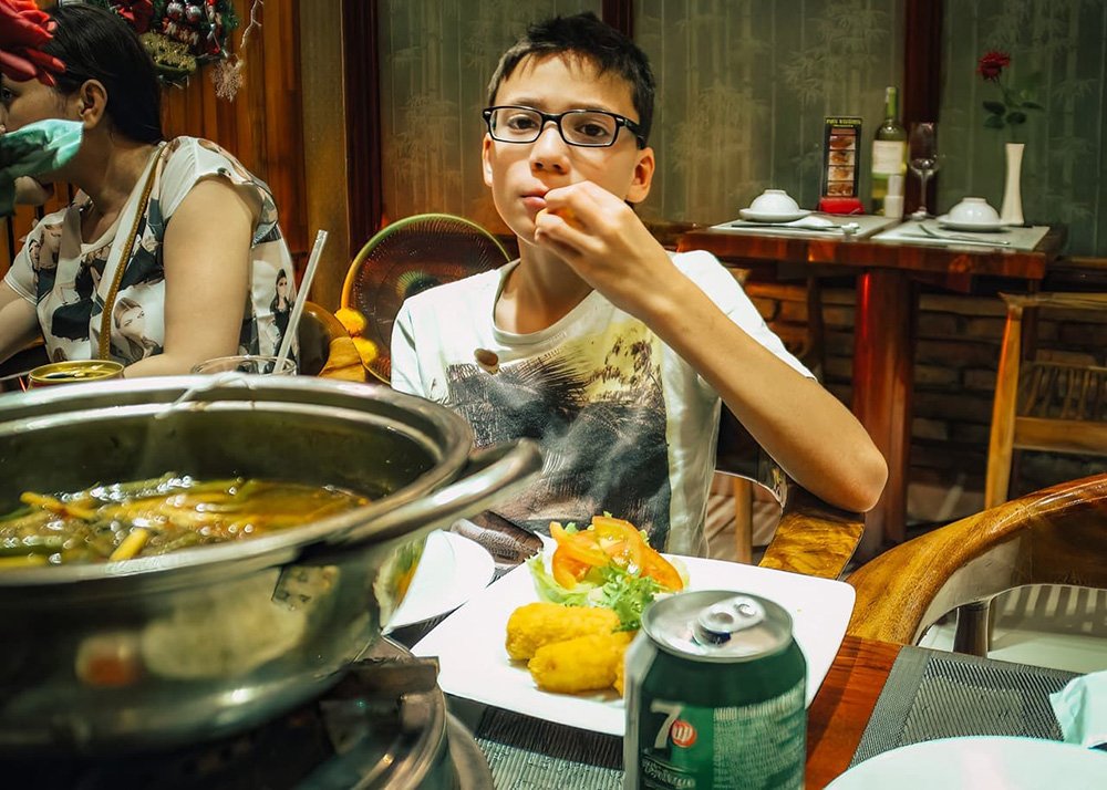 A boy eating a spring roll with a large hot pot in front of him and a plate with spring rolls and a 7up.