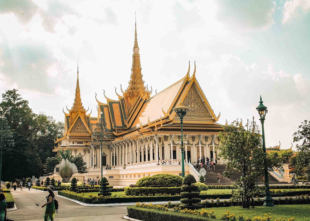 A magnificent golden-roofed building at the Royal Palace in Phnom Penh, Cambodia, on a partly cloudy day. The ornate architecture features spires and intricate designs, surrounded by lush gardens and visitors exploring the grounds.