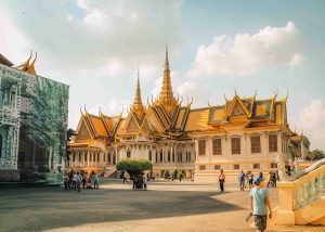 The Royal Palace in Phnom Penh, Cambodia, showcasing its grand golden rooftops and multiple spires against a bright blue sky. Tourists are walking around the expansive courtyard, capturing the historical beauty of the palace.