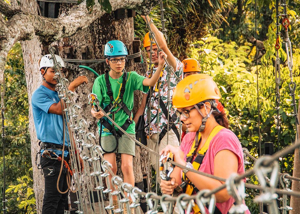 A family zip-lining adventure at the Flight of the Gibbon in Angkor Park, Cambodia, showing participants wearing helmets and harnesses. The sunny day and lush forest canopy make for an exciting outdoor activity.