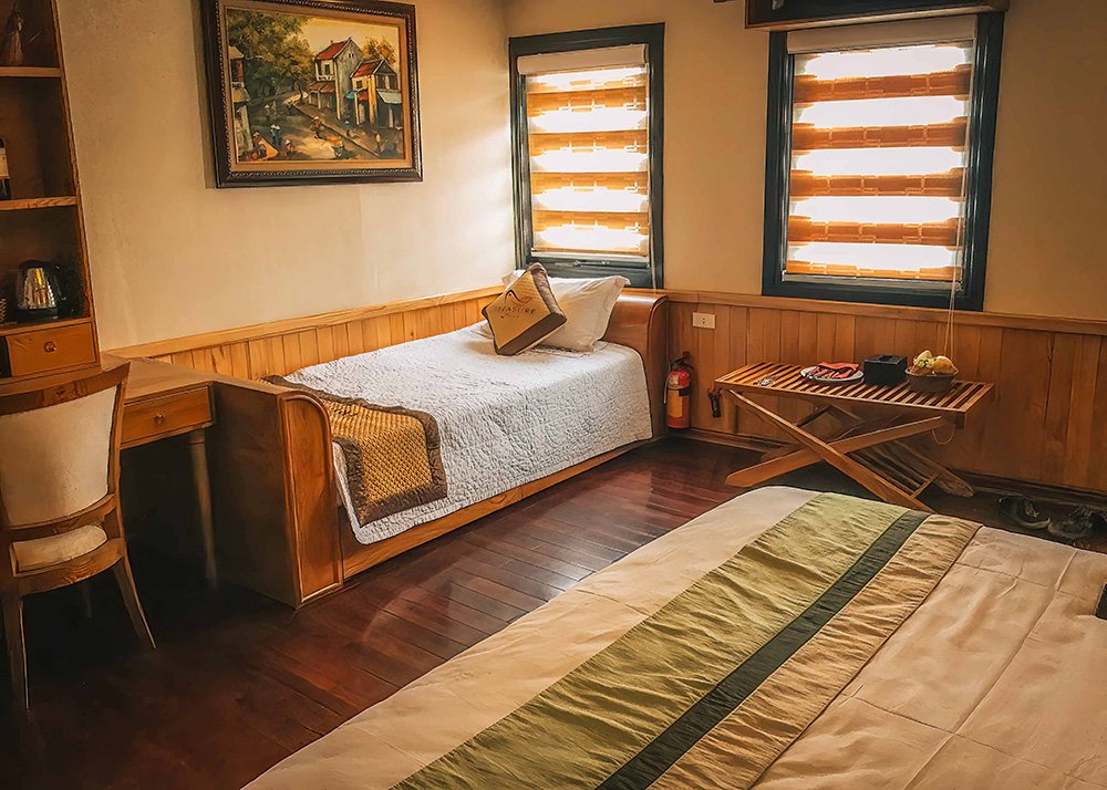 Cozy hotel room with single bed and desk: A warmly lit hotel room featuring a single bed with a white quilt, a desk and chair, and two windows with blinds partially closed.