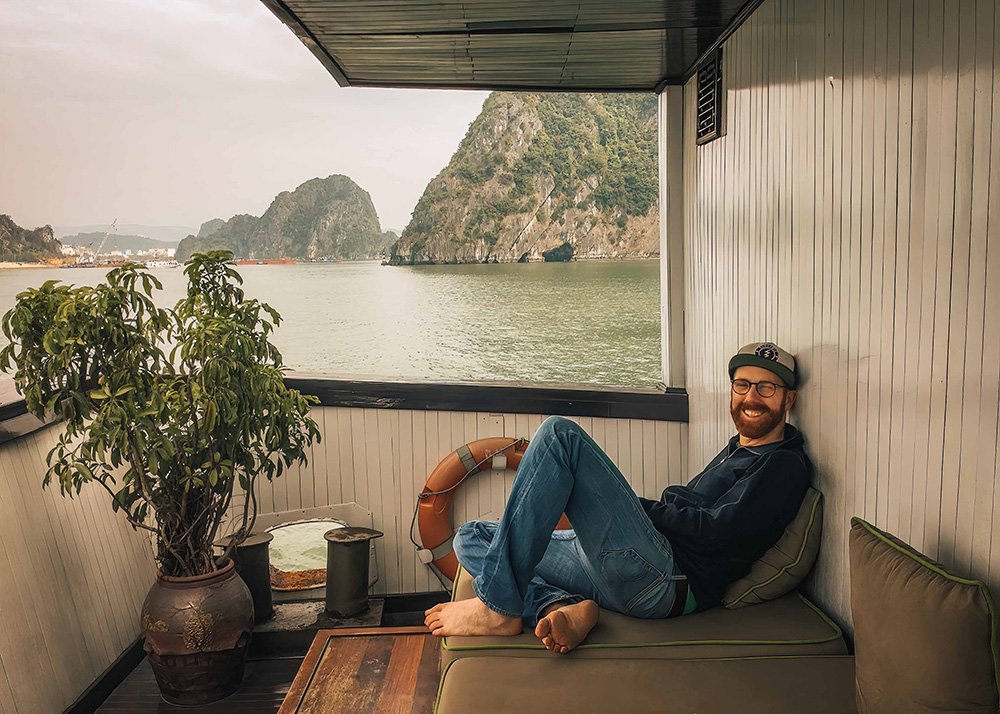Man relaxing on a boat balcony in Halong Bay: A smiling man with a beard and glasses relaxes on a cushioned bench on a boat balcony, overlooking the scenic limestone cliffs of Halong Bay.