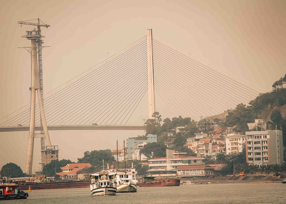 View of a suspension bridge and boats in Halong Bay: A view of a suspension bridge spanning over Halong Bay, with boats and buildings along the shoreline, captured in a hazy atmosphere.