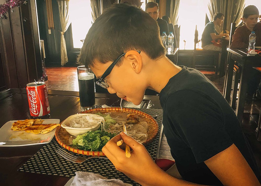Boy eating Vietnamese food on a boat: A young boy enjoying a traditional Vietnamese meal on a boat, with a bowl of noodles, greens, and a can of Coca-Cola on the table.