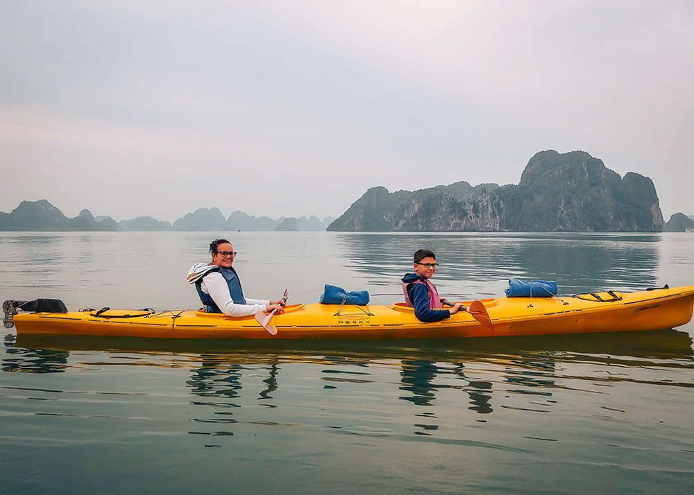 Mother and son kayaking in Halong Bay: A mother and her son paddle a yellow kayak in the calm waters of Halong Bay, with misty islands in the distance.