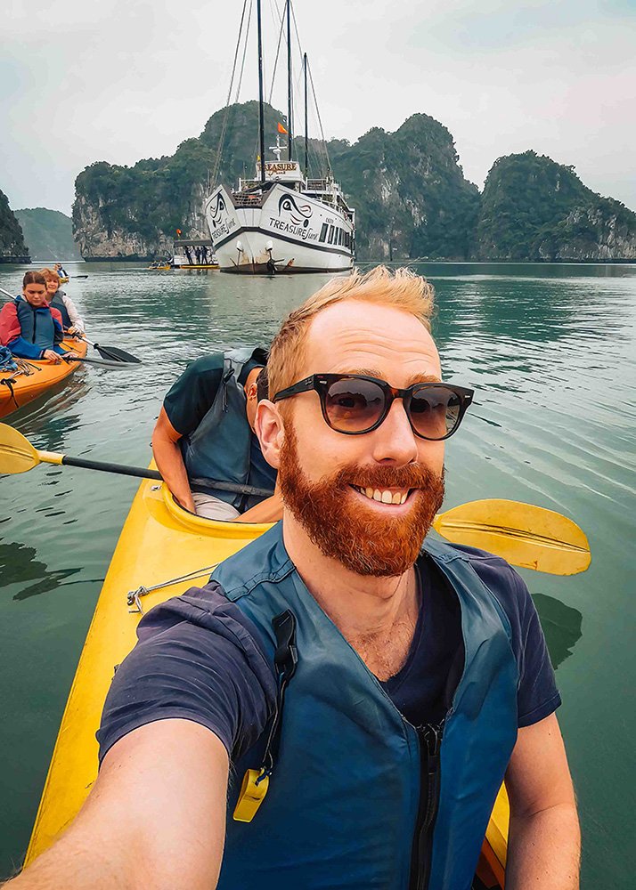 Father taking a selfie while kayaking in Halong Bay: A smiling father wearing sunglasses takes a selfie from his yellow kayak, with a boat and other kayakers visible in the background.