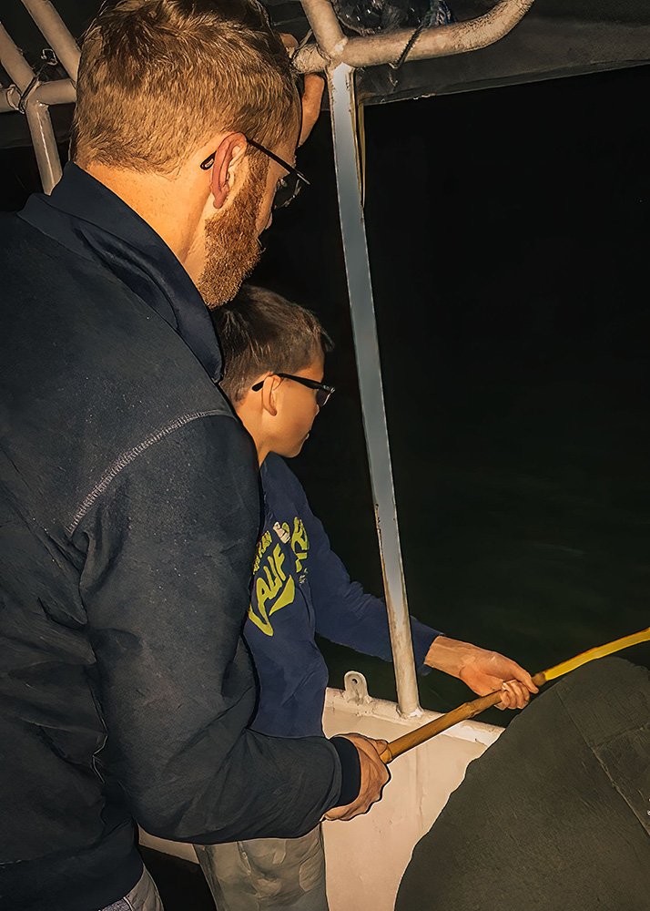 Father and son night fishing on a boat: A father and his son fishing from the side of a boat at night, both focused on their activity under dim lighting.