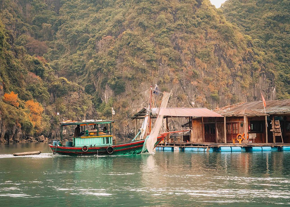 Fishing boat near floating village in Halong Bay: A green and red fishing boat docked near rustic houses of a floating village in Halong Bay, with limestone cliffs in the background.