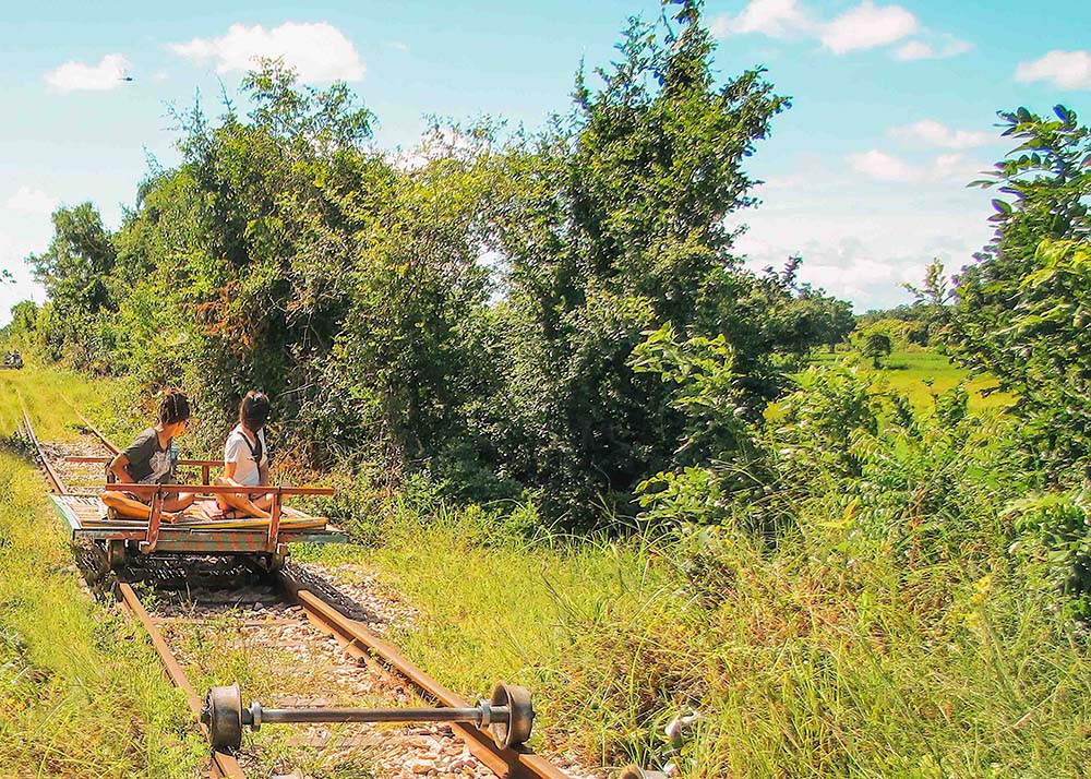 Two women riding a bamboo train through the lush countryside of Battambang, Cambodia, on a sunny day. The open-air ride offers a unique perspective of the surrounding greenery.
