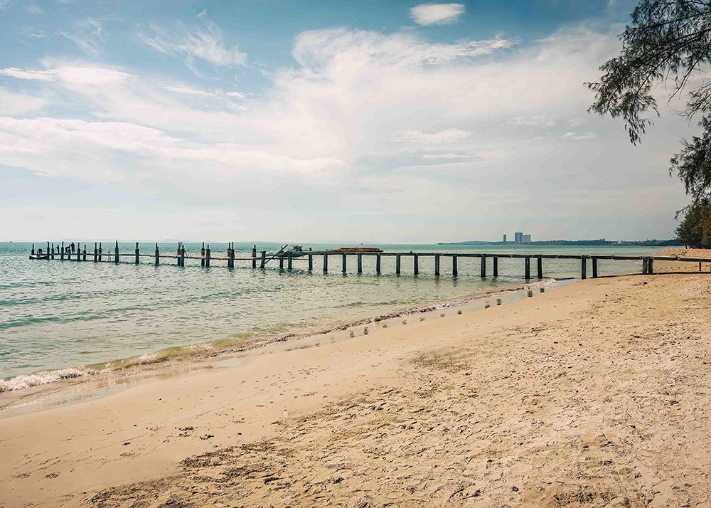 A quiet beach in Sihanoukville, Cambodia, with a long wooden pier stretching into the calm sea under a partly cloudy sky. The empty shoreline and gentle waves create a peaceful atmosphere.