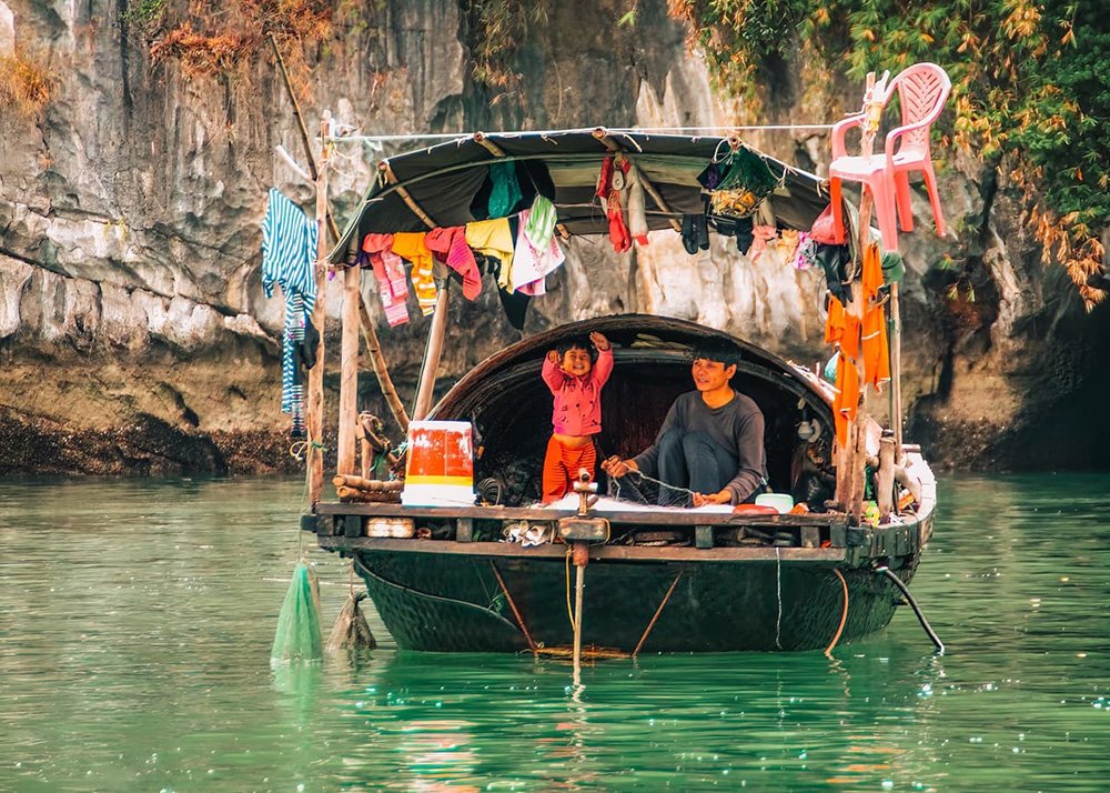 Local family on a boat in Halong Bay: A local man and a child on a small boat in Halong Bay, with colorful clothes hanging to dry and limestone cliffs in the background.